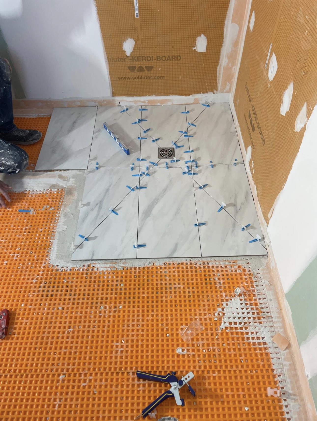 Marble-look shower floor tile being set on Schluter KERDI around the drain, with orange uncoupling membrane still exposed