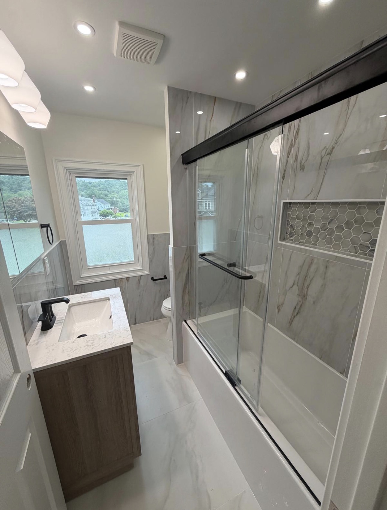 Finished bathroom with light-oak vanity, marble-look walls, and a sliding glass tub and shower with hexagonal niche